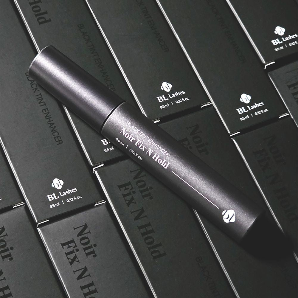 BL Lashes Black Tint Enhancer Noir Fix N Hold tube resting diagonally across multiple of its black packaging boxes.