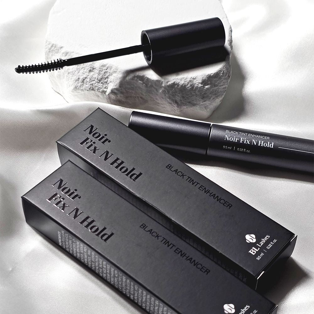 BL Lashes Black Tint Enhancer Noir Fix N Hold tube and two packaging boxes on white silk fabric, with its opened curved wand resting on a textured white stone.
