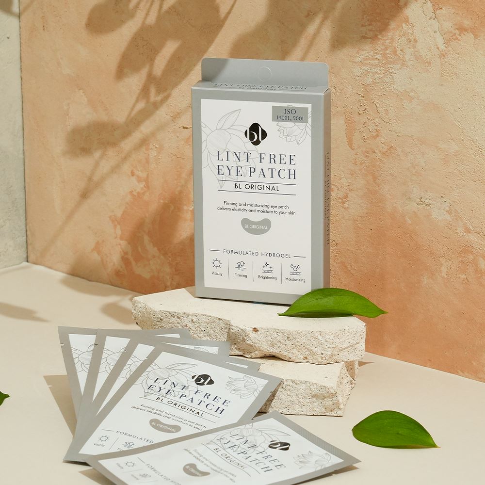 BL Lashes Lint Free Eye Patch “BL Original” hydrogel eye patch box displayed on stacked textured stone blocks with several individual gray sachets spread in front. The packaging reads “Lint Free Eye Patch BL Original” and “Formulated Hydrogel,” with icons highlighting vitality, firming, brightening, and moisturizing benefits. Warm natural light, a textured beige background, and green leaves create a clean beauty product composition for under-eye care during eyelash extension treatments.