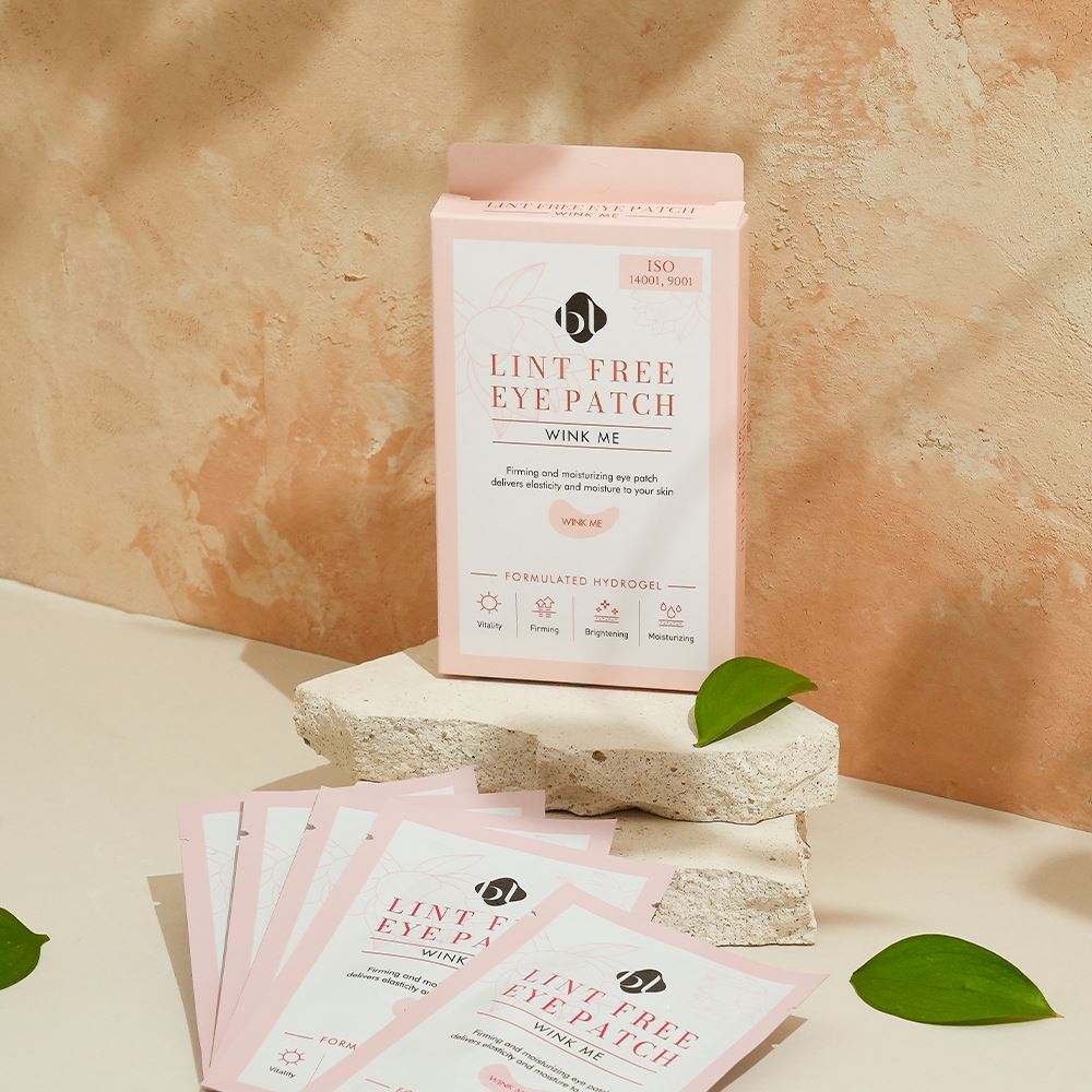 BL Lashes Lint Free Eye Patch “Wink Me” hydrogel eye patch box displayed on a textured stone platform with several individual pink sachets spread in front. The packaging reads “Lint Free Eye Patch Wink Me” and “Formulated Hydrogel,” with icons highlighting vitality, firming, brightening, and moisturizing benefits. The warm-toned background and green leaves create a natural beauty product composition designed for under-eye care during eyelash extension treatments.