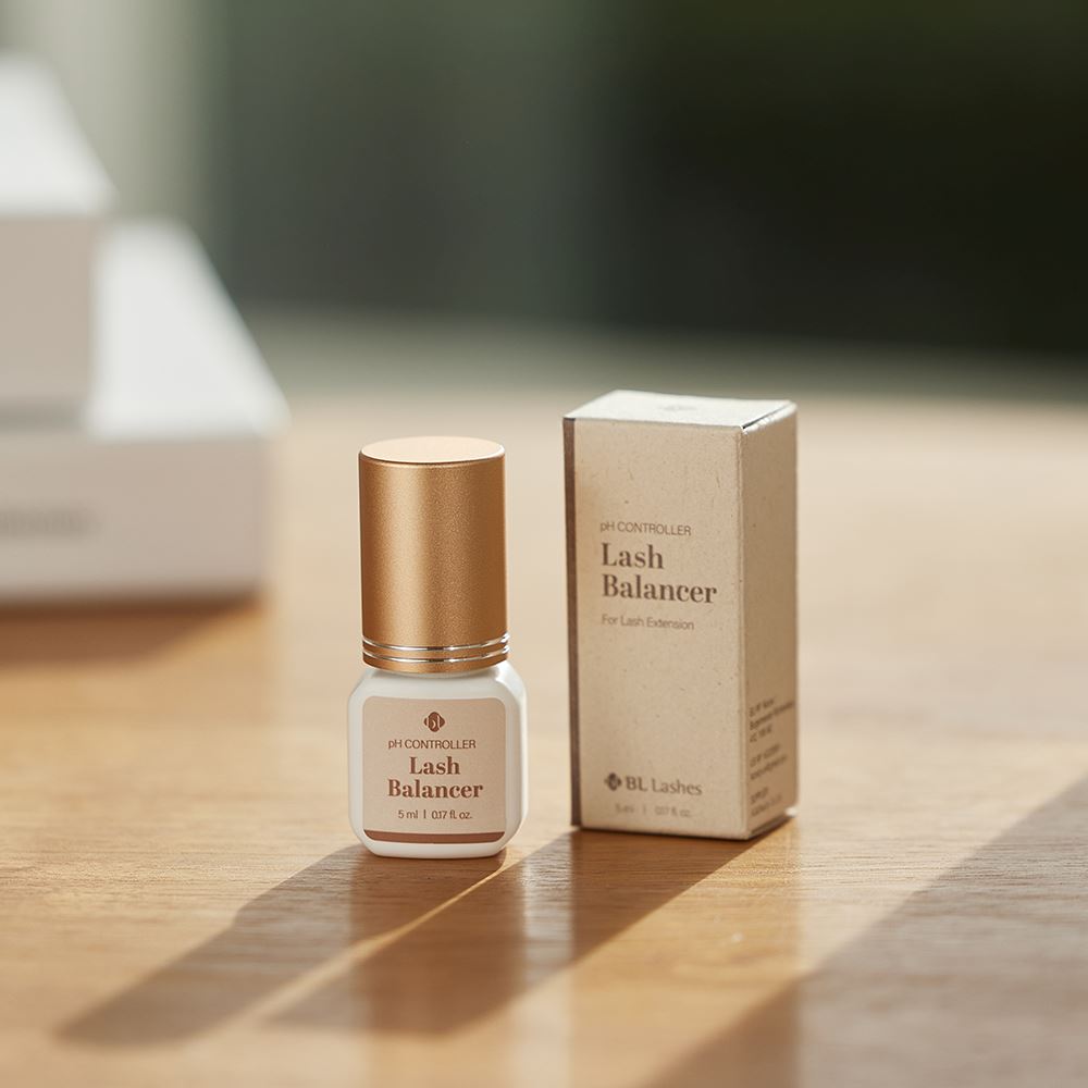 BL Lashes Lash Balancer pH Controller in a small frosted glass bottle with a metallic gold cap displayed beside its beige product box on a wooden surface. The label reads “pH Controller Lash Balancer” with the volume “5 ml / 0.17 fl. oz.” Warm natural light casts soft shadows, creating a lifestyle-style product presentation that highlights the compact professional solution designed for pH balancing during eyelash extension preparation.