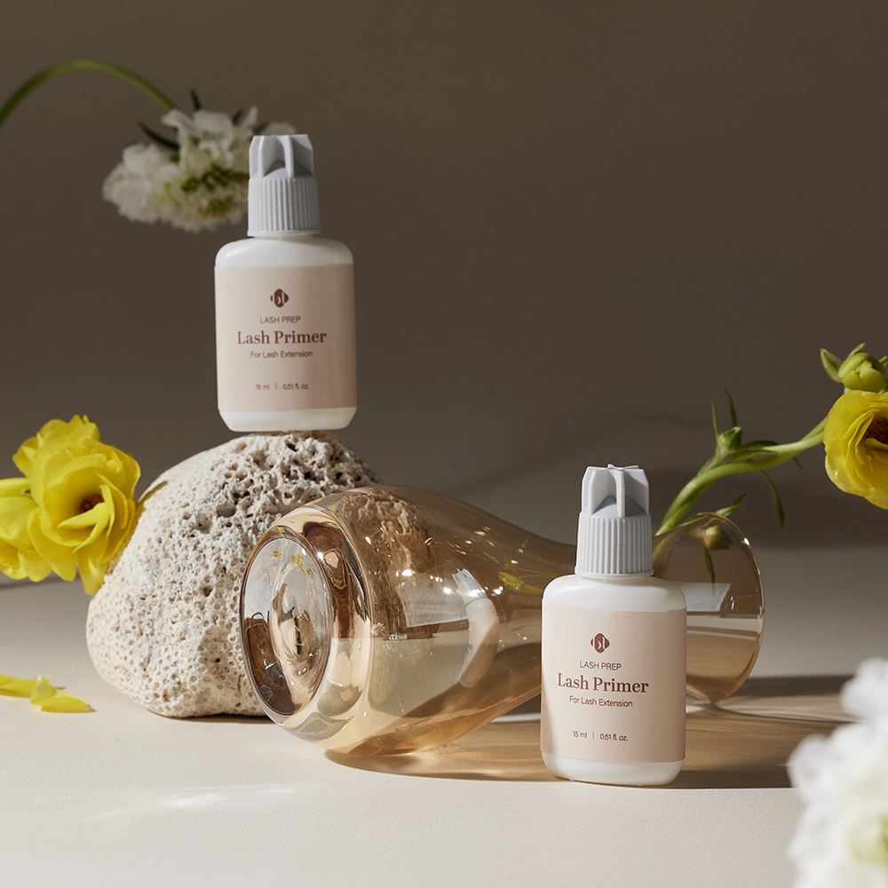 Two BL Lashes Lash Prep Lash Primer 15ml bottles arranged with a textured stone, a tipped amber glass vase, and delicate yellow and white flowers.