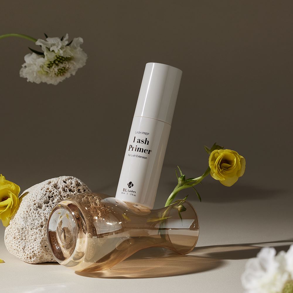 BL Lashes Lash Prep Lash Primer bottle resting on a tipped amber glass vase, arranged with a textured stone and delicate yellow and white flowers.