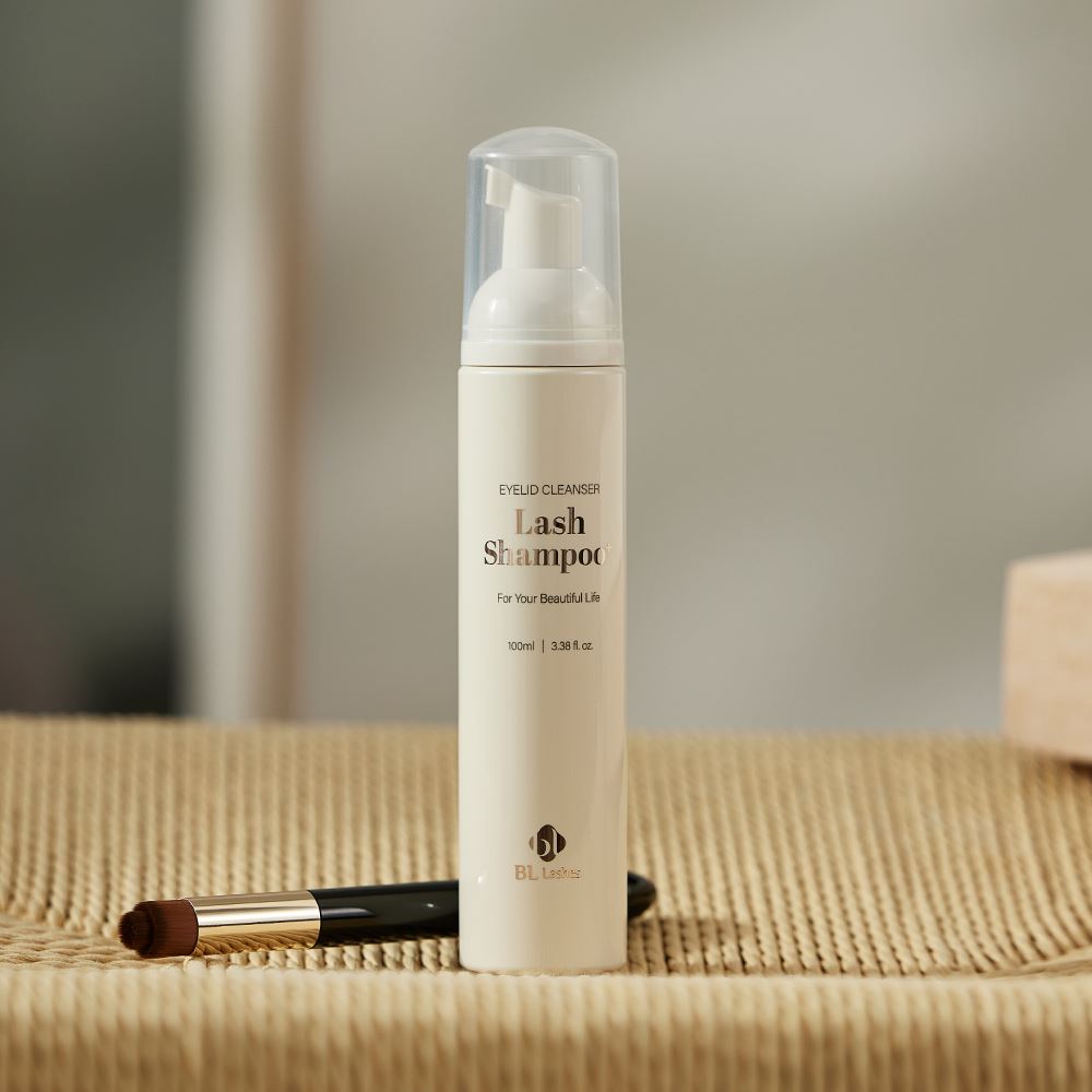BL Lashes Lash Shampoo eyelid cleanser in a minimalist beige bottle with a foaming pump and transparent cap stands on a textured woven mat. The packaging features rose-gold lettering reading “Lash Shampoo,” along with “Eyelid Cleanser,” “For Your Beautiful Life,” and “100 ml / 3.38 fl. oz.” A small cleansing brush with brown bristles and a black handle lies beside the bottle, highlighting its use for eyelash extension cleansing and gentle lash care in a warm, softly lit setting.