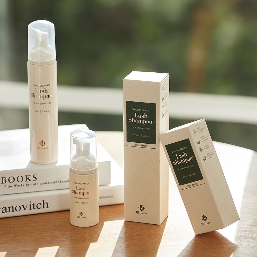 BL Lashes Lash Shampoo eyelid cleanser bottles in beige foaming pump containers are displayed on stacked books beside their product boxes on a wooden table. The packaging features elegant lettering reading ?œLash Shampoo,???œEyelid Cleanser,??and ?œFor Your Beautiful Life,??with sizes including 100 ml / 3.38 fl. oz. and 50 ml / 1.69 fl. oz. Soft natural light and a blurred green background create a warm, lifestyle-style presentation highlighting eyelash extension cleansing and lash care products.