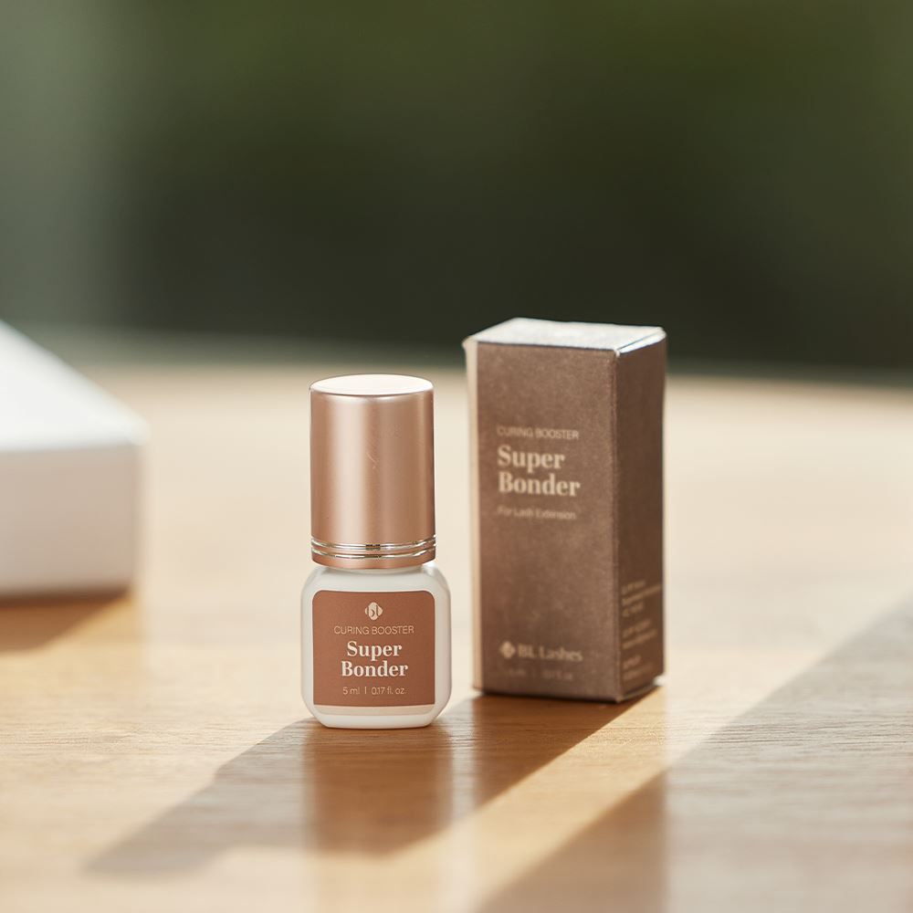 BL Lashes Super Bonder curing booster for lash extensions in a small frosted glass bottle with a metallic rose gold cap displayed beside its brown product box on a wooden surface. The label reads “Curing Booster Super Bonder” with the volume “5 ml / 0.17 fl. oz.” Warm natural light casts soft shadows across the table, creating a lifestyle-style beauty product composition that highlights a compact professional solution designed to accelerate adhesive curing for eyelash extensions.