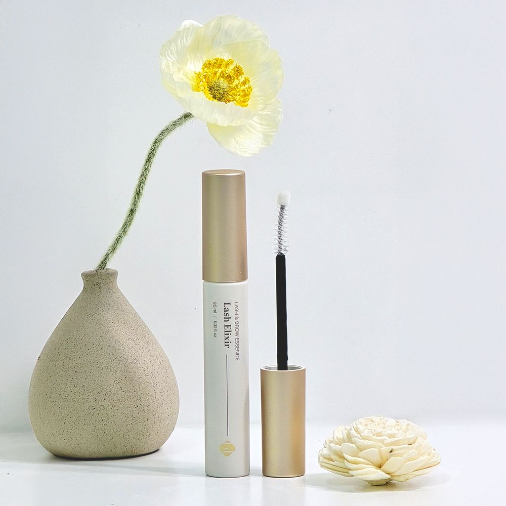 BL Lashes Lash Elixir tube and opened wand arranged with a pale yellow flower in a small vase and a white decorative flower.